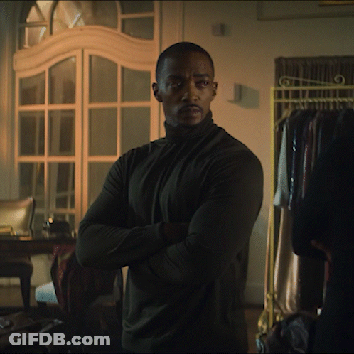 A Man In A Black Turtleneck Stands With His Arms Crossed In A Dark Room GIF
