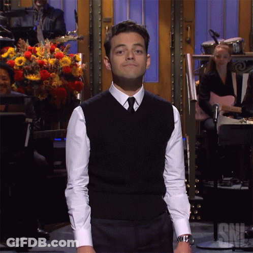 A Man In A Black Vest And White Shirt Stands In Front Of A Snl Sign GIF