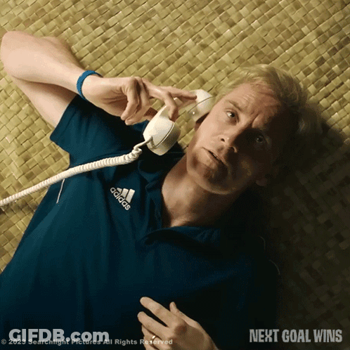 A Man In A Blue Adidas Shirt Is Laying On The Floor Talking On A Telephone GIF