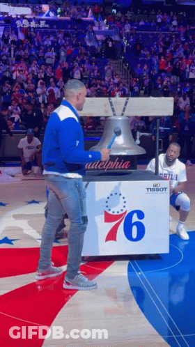 A Man In A Blue And White Jacket Is Standing Next To A Bell With The Number 76 On It GIF