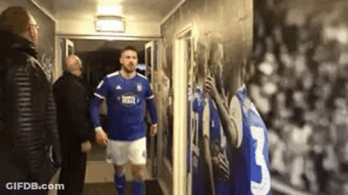 A Man In A Blue And White Soccer Uniform Is Walking Down A Hallway With Two Men . GIF