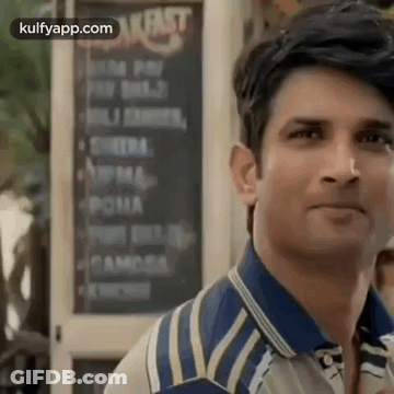 A Man In A Blue And White Striped Shirt Is Standing In Front Of A Restaurant . GIF