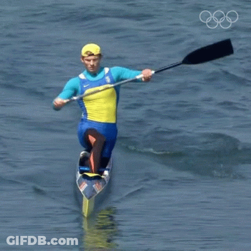 A Man In A Blue And Yellow Outfit Is Paddling A Canoe In The Water With The Olympic Rings In The Background GIF