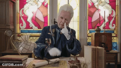 A Man In A Blue Coat And White Gloves Is Sitting At A Desk With A Stained Glass Window In The Background GIF
