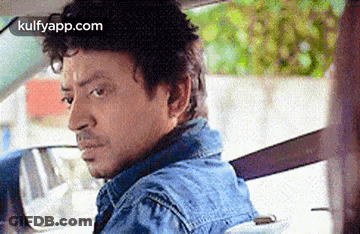 A Man In A Blue Denim Jacket Is Driving A Car And Looking At The Rear View Mirror . GIF