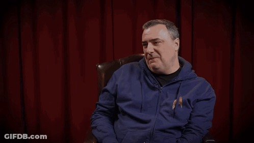 A Man In A Blue Hoodie Sits In Front Of A Red Curtain With The Words " Wide Beam That On Kil " Behind Him GIF