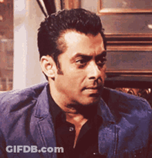 A Man In A Blue Jacket And Black Shirt Is Sitting In A Chair . GIF