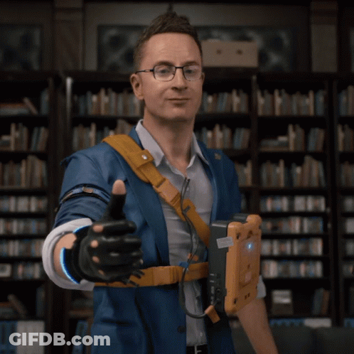 A Man In A Blue Jacket Giving A Thumbs Up In Front Of A Bookshelf GIF