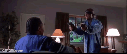 A Man In A Blue Jacket Is Pointing A Gun At Another Man Sitting On A Couch . GIF