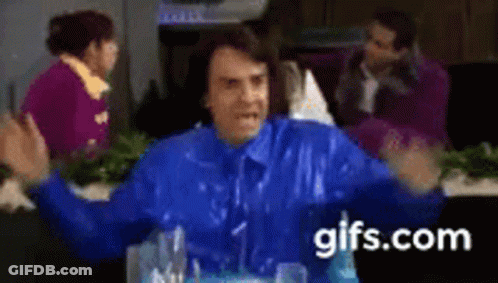 A Man In A Blue Jacket Is Sitting At A Table With His Arms Outstretched In Front Of A Group Of People . GIF