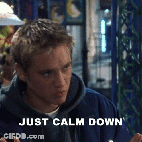 A Man In A Blue Jacket Is Sitting At A Table With The Words `` Just Calm Down '' Written On The Screen . GIF