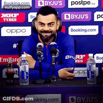 A Man In A Blue Jacket Is Sitting In Front Of A Microphone Holding A Bottle Of Water . GIF
