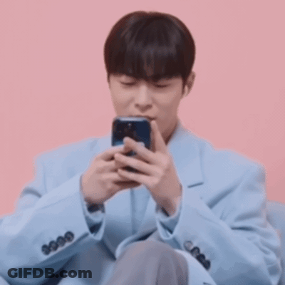 A Man In A Blue Jacket Is Sitting On A Pink Couch Looking At His Cell Phone . GIF