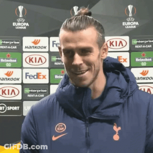 A Man In A Blue Jacket Is Smiling In Front Of A Europa League Banner GIF