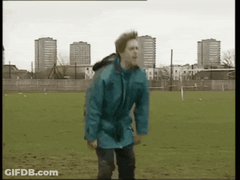 A Man In A Blue Jacket Is Standing In A Field With Tall Buildings In The Background GIF