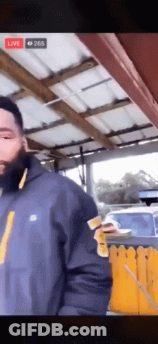 A Man In A Blue Jacket Is Standing In Front Of A Yellow Fence While Holding A Piece Of Paper . GIF