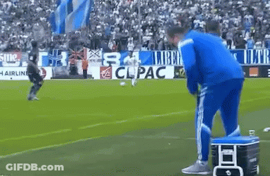 A Man In A Blue Jacket Is Standing On A Soccer Field Next To A Cooler .. GIF