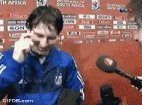 A Man In A Blue Jacket Is Talking Into A Microphone While Another Man Looks On GIF