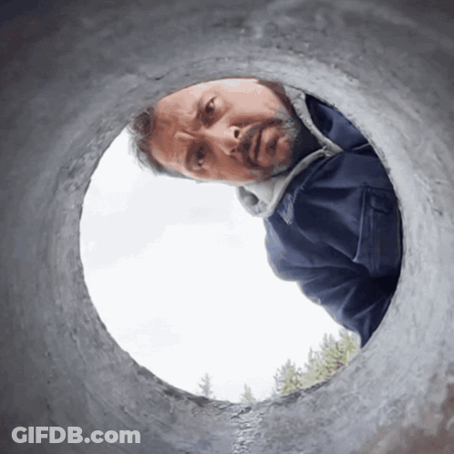 A Man In A Blue Jacket Looks Through A Hole In A Pipe GIF