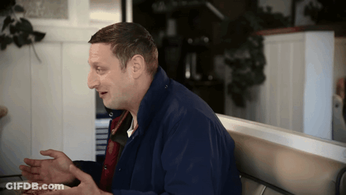 A Man In A Blue Jacket Sits At A Table With His Hands Out GIF