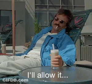 A Man In A Blue Jacket Sits In A Chair Holding A Drink And A Cigarette GIF