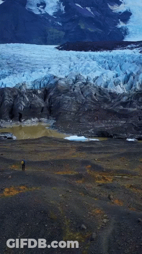 A Man In A Blue Jacket Stands In Front Of A Glacier GIF