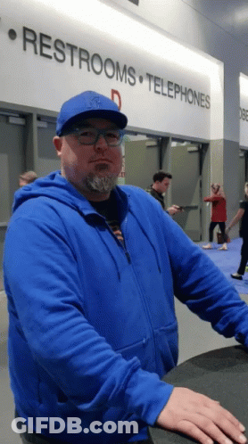 A Man In A Blue Jacket Stands In Front Of A Sign That Says Restrooms And Telephones GIF