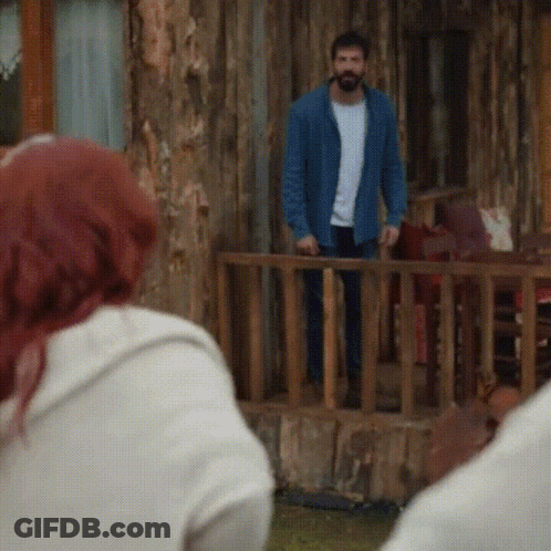 A Man In A Blue Jacket Stands In Front Of A Wooden House GIF