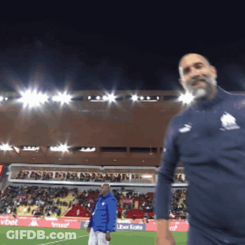 A Man In A Blue Jacket Stands On A Soccer Field In Front Of An Uber Eats Advertisement GIF