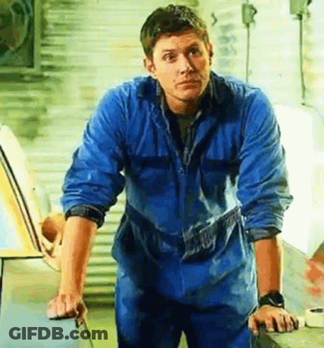A Man In A Blue Jumpsuit Is Standing In Front Of A Car . GIF