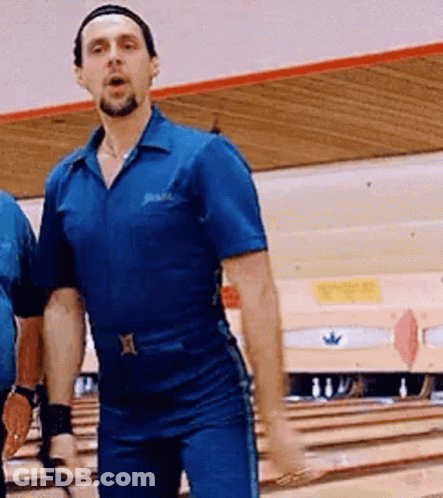 A Man In A Blue Jumpsuit Is Standing On A Bowling Alley With His Hands In The Air . GIF