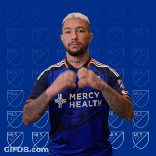 A Man In A Blue Mercy Health Jersey Makes A Heart Shape With His Hands GIF