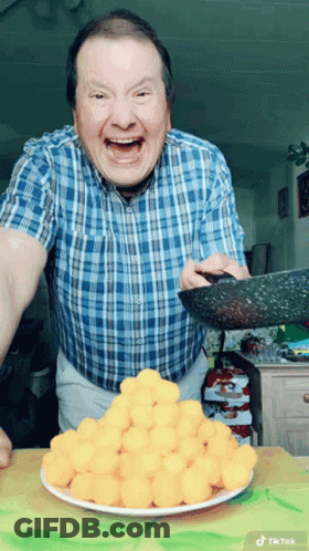 A Man In A Blue Plaid Shirt Is Holding A Pan Over A Plate Of Cheese Balls GIF