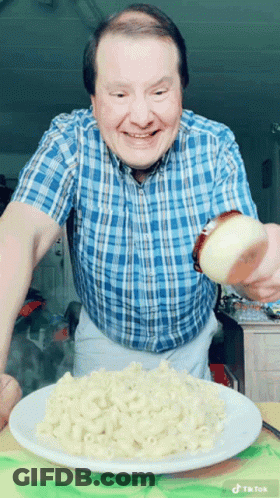 A Man In A Blue Plaid Shirt Is Pouring A Slice Of Onion On A Plate Of Macaroni And Cheese GIF
