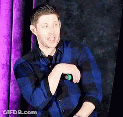 A Man In A Blue Plaid Shirt Is Standing In Front Of A Purple Curtain And Talking Into A Microphone GIF