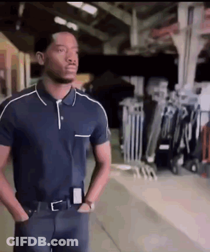 A Man In A Blue Polo Shirt Is Standing In A Warehouse With His Hands In His Pockets GIF