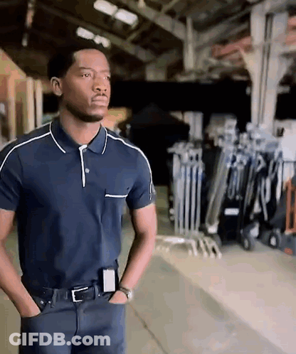 A Man In A Blue Polo Shirt Is Standing With His Hands In His Pockets In A Warehouse . GIF