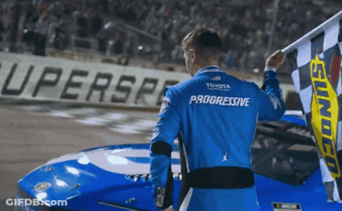 A Man In A Blue Progressive Shirt Holds A Checkered Flag GIF