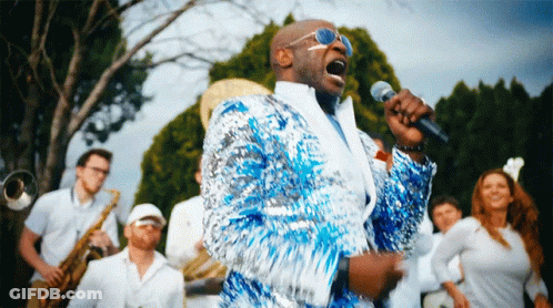 A Man In A Blue Sequined Jacket Sings Into A Microphone In Front Of A Band GIF