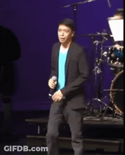 A Man In A Blue Shirt And A Black Jacket Is Singing Into A Microphone On A Stage GIF