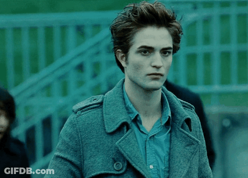 A Man In A Blue Shirt And A Grey Coat Stands In Front Of A Green Fence GIF