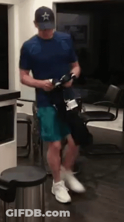 A Man In A Blue Shirt And A Hat Is Standing In A Room Holding A Bottle . GIF