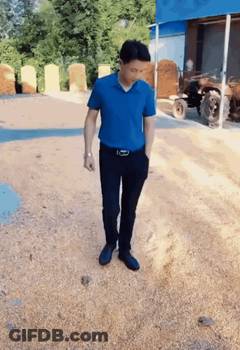 A Man In A Blue Shirt And Black Pants Is Standing On A Gravel Road GIF