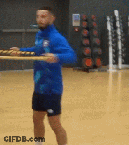 A Man In A Blue Shirt And Black Shorts Is Holding A Wooden Plank . GIF