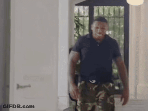 A Man In A Blue Shirt And Camo Pants Is Standing In A Hallway . GIF