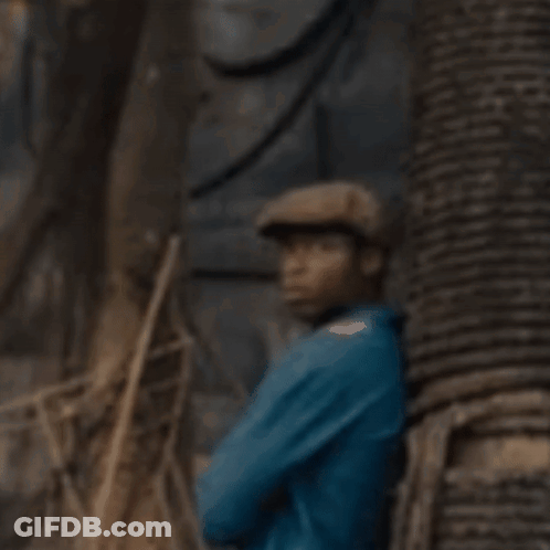 A Man In A Blue Shirt And Hat Is Standing Next To A Tree . GIF