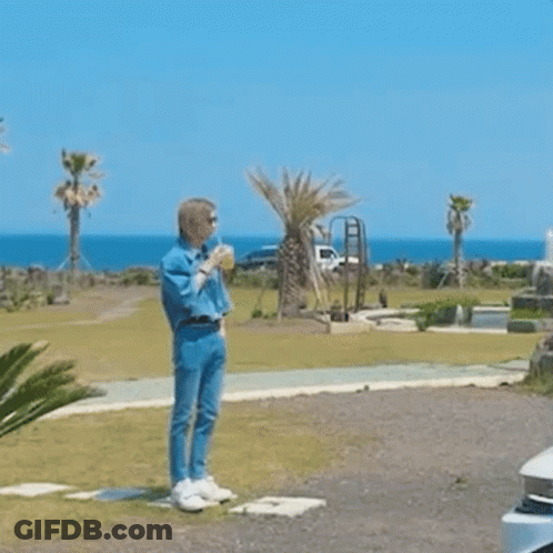 A Man In A Blue Shirt And Jeans Is Standing In A Field Drinking A Drink . GIF