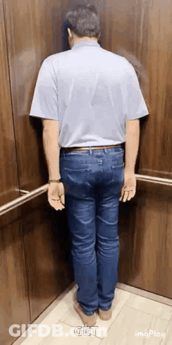 A Man In A Blue Shirt And Jeans Is Standing In An Elevator . GIF