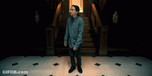 A Man In A Blue Shirt And Jeans Is Standing In Front Of A Set Of Stairs GIF