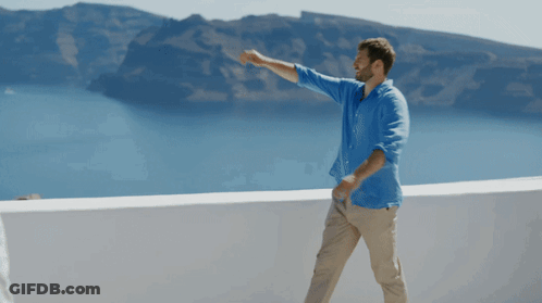 A Man In A Blue Shirt And Khaki Pants Stands On A Balcony Overlooking The Ocean GIF